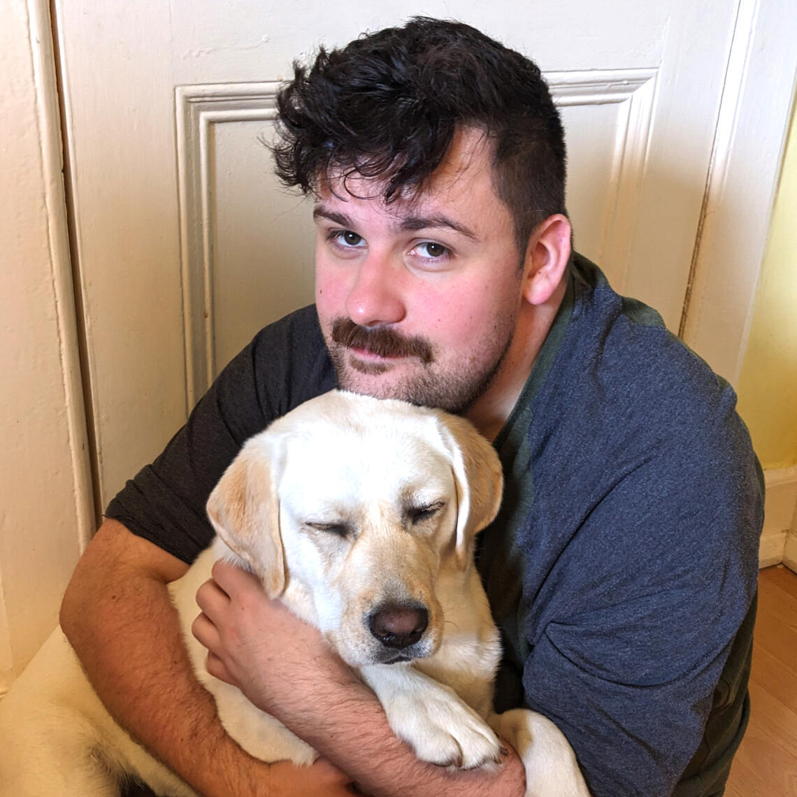 A photo of Brian Tyrrell. He has short black hair, an upper lip moustache, and stubble. He is holding a white labrador in his arms.